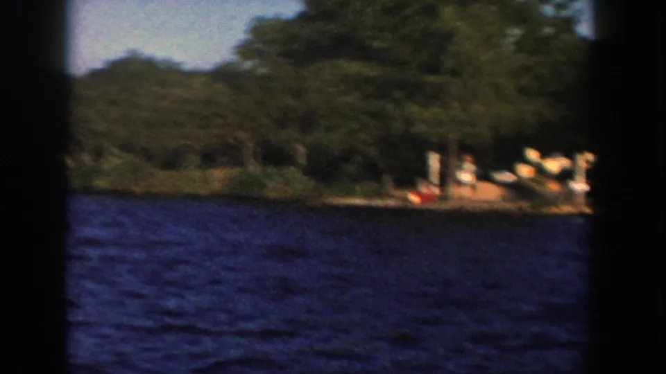 1958 Wisconsin 8mm view from a boat, with choppy blue water, dense trees, and a small dock or shore landing at right.