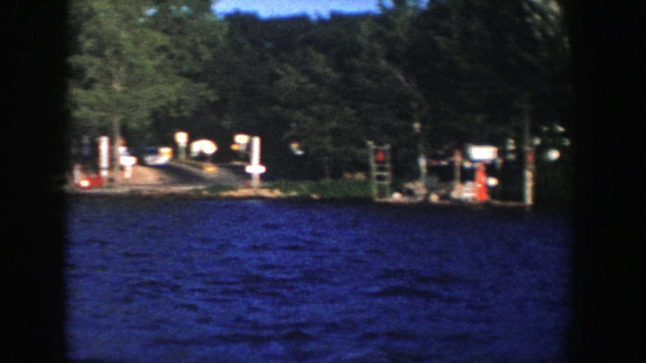 1958 Wisconsin home-movie frame showing lake water in front of a wooded dock area with posts, signs, and a small structure.