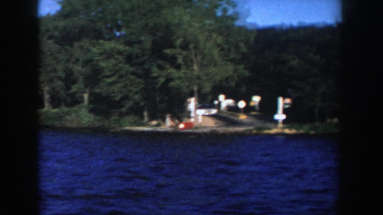 1958 Wisconsin 8mm frame with rippled lake water, dark boat framing, trees, dockside posts, and small pale signs on shore.