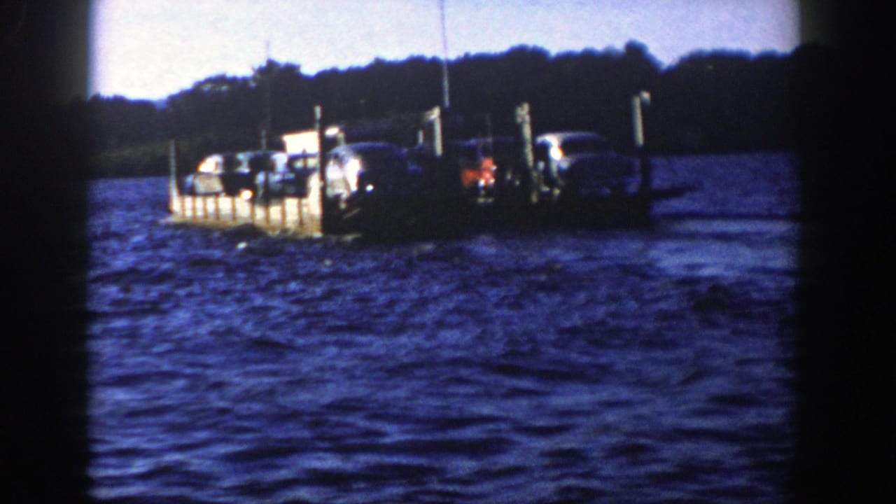 1958 Wisconsin home-movie frame of a low car ferry crossing a lake, with upright posts, automobiles, and distant trees visible.