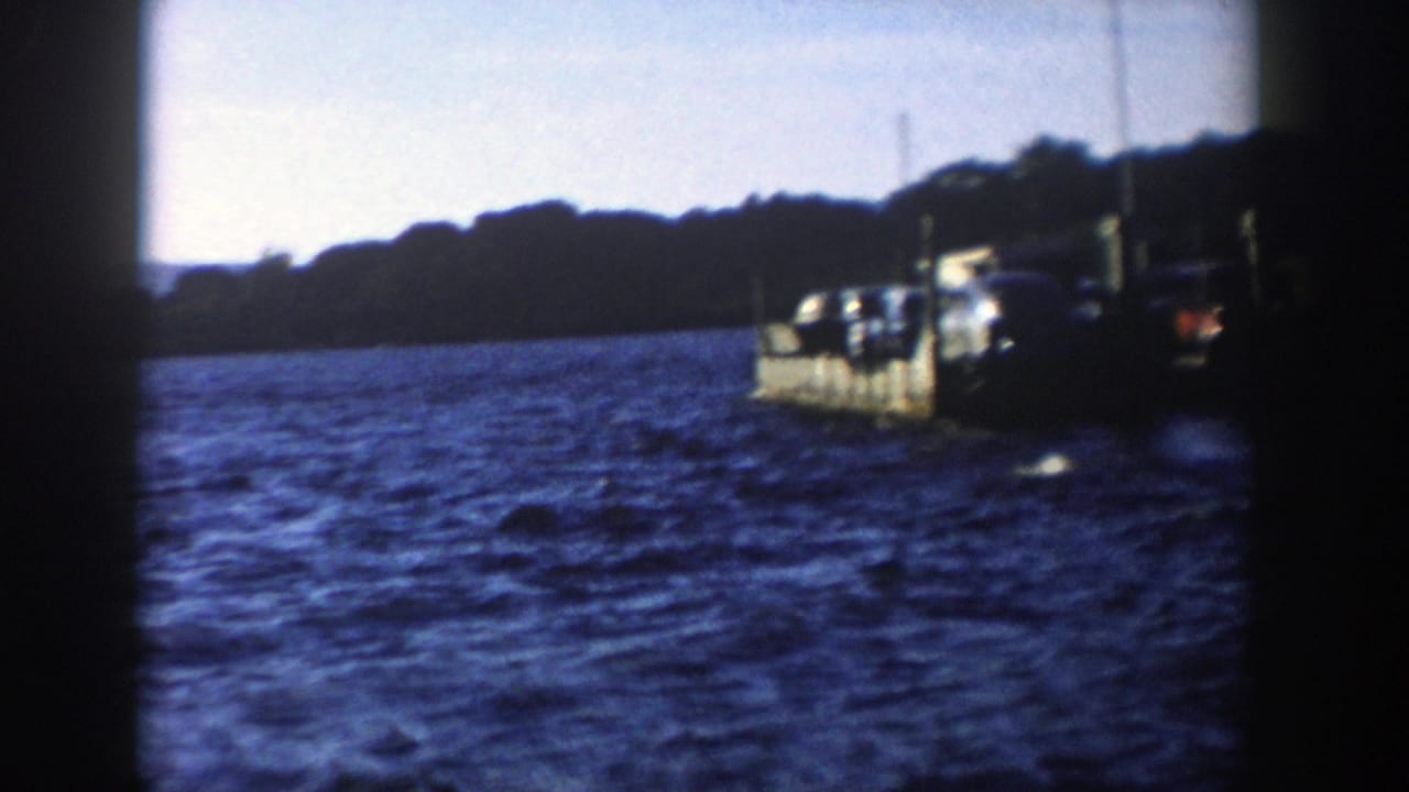 Soft 8mm Wisconsin lake view from 1958, showing a car ferry partly framed by dark film edges and a wooded shore behind it.