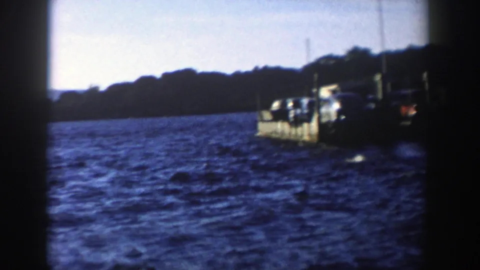 Soft 8mm Wisconsin lake view from 1958, showing a car ferry partly framed by dark film edges and a wooded shore behind it.