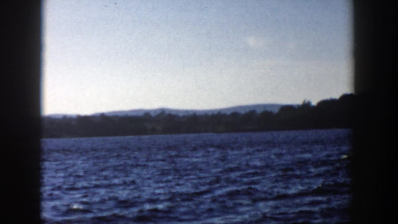 1958 Wisconsin 8mm frame opening to a wider lake view after the ferry passes, with distant hills and shoreline under a pale sky.