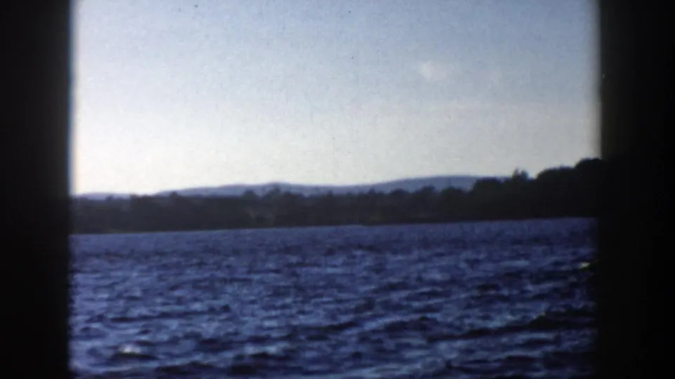 1958 Wisconsin 8mm frame opening to a wider lake view after the ferry passes, with distant hills and shoreline under a pale sky.