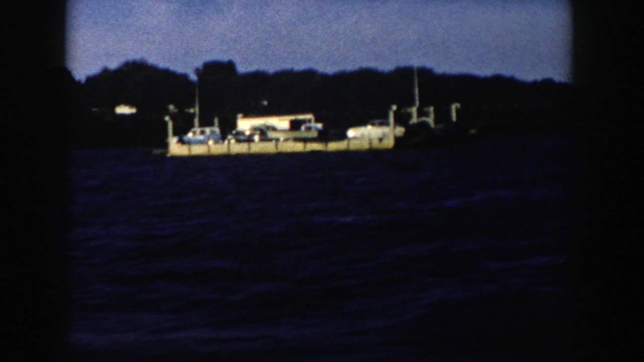 1958 Wisconsin 8mm frame showing a light-colored barge or ferry crossing rough blue water below a dark wooded shoreline.