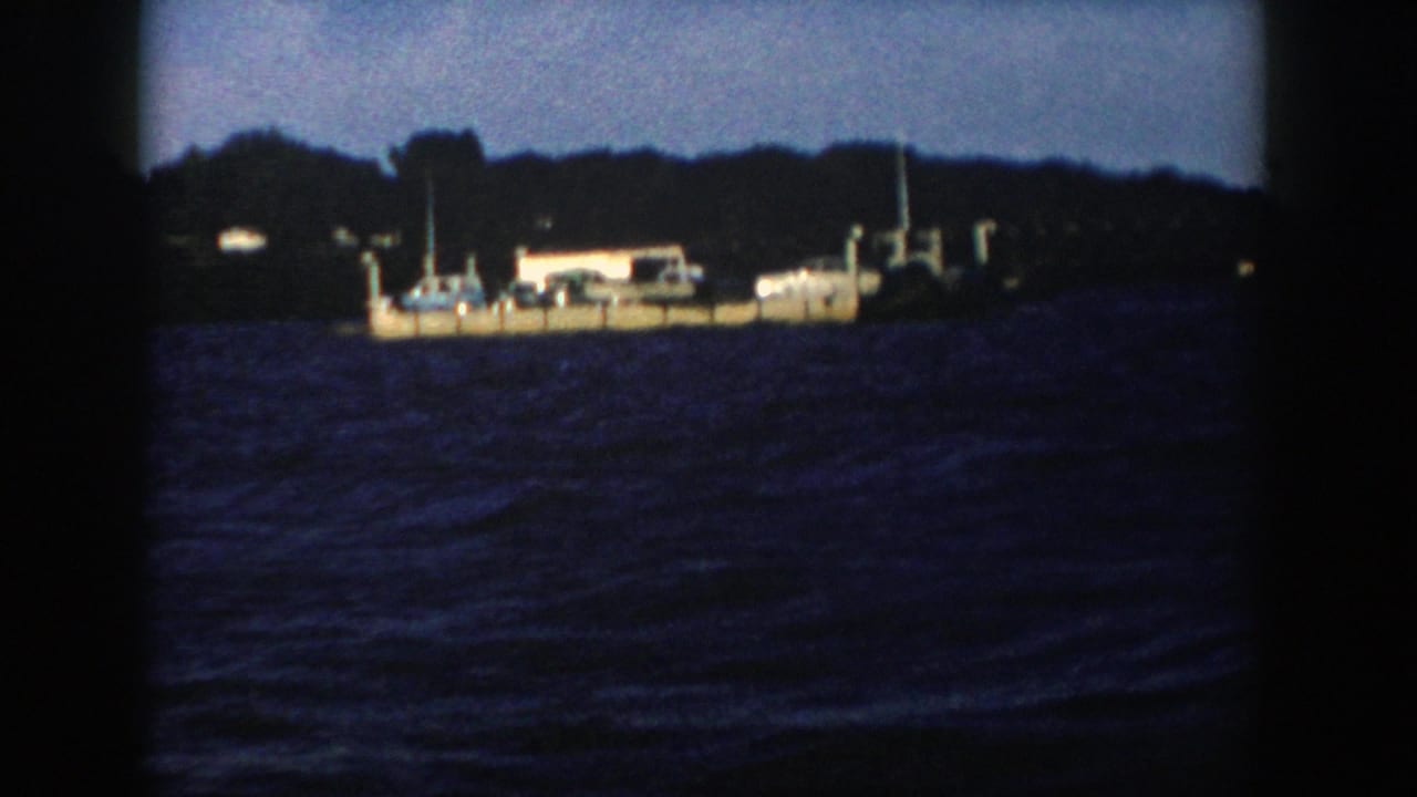 1958 Wisconsin home-movie frame of a barge or ferry viewed from the water, with wooded shore silhouetted behind it.