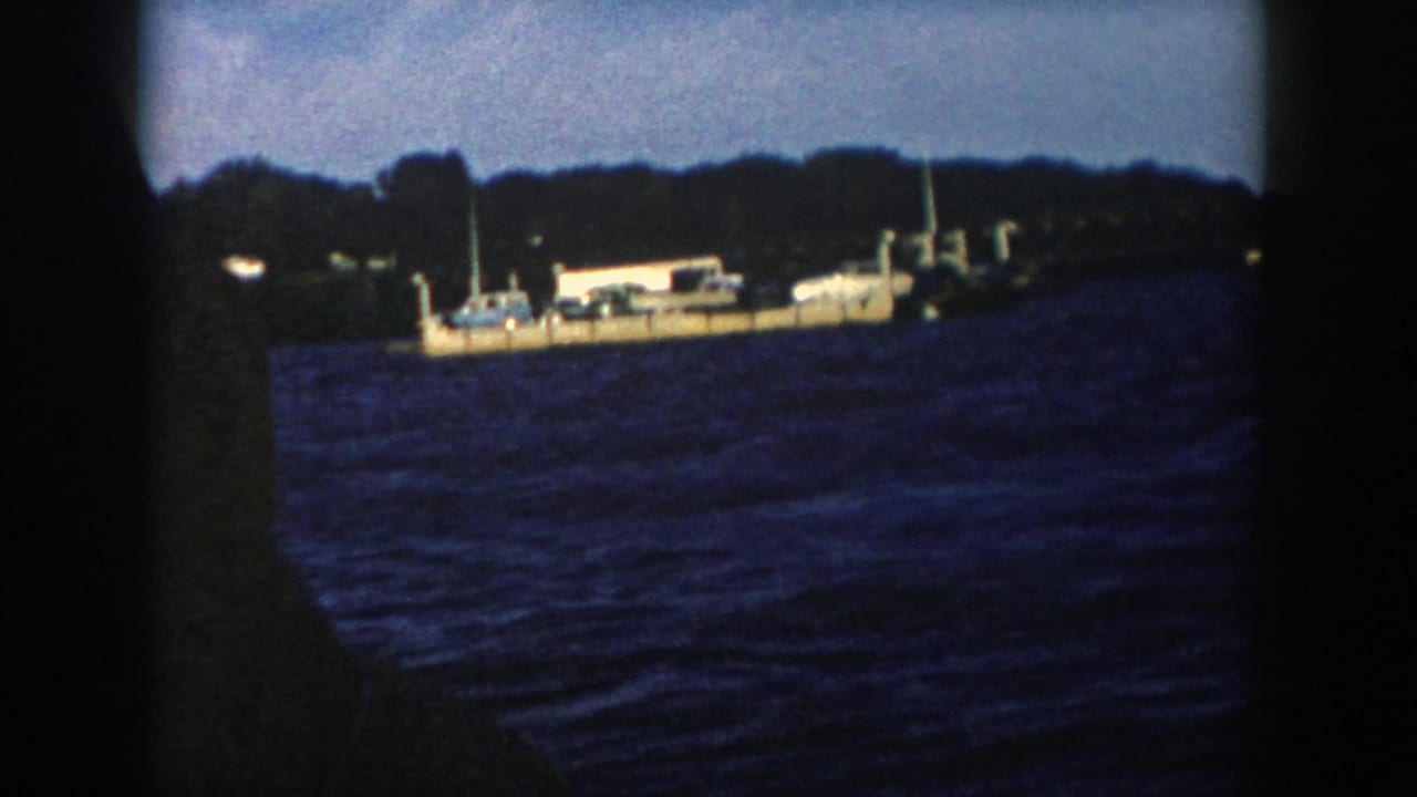 1958 Wisconsin home-movie frame showing a distant barge or ferry on turbulent blue water beneath a silhouetted tree line.