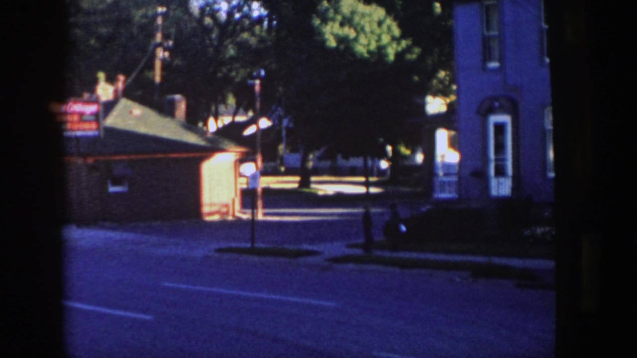 Small roadside building with a glowing sign, house facade, street, trees, and a pedestrian at dusk.