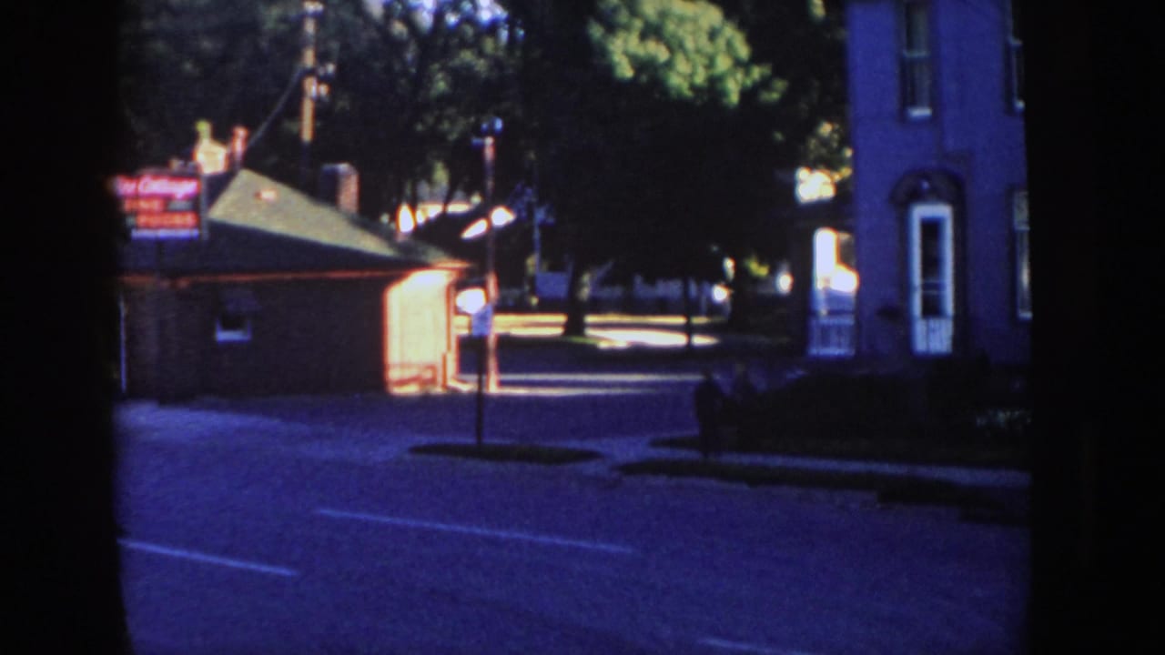 1958 Wisconsin home-movie frame of a roadside sign, small building, shaded house, street, trees, and a distant person on the sidewalk.