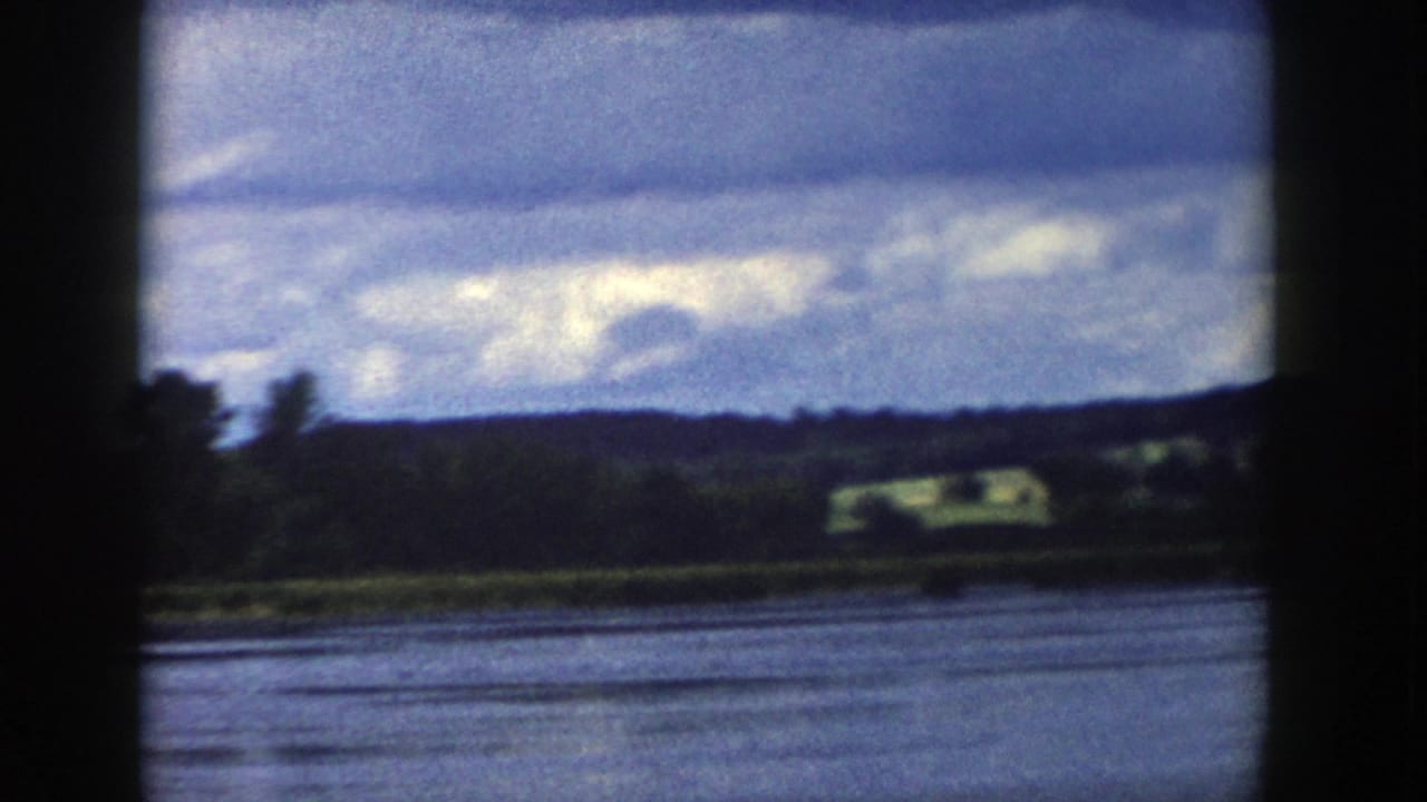 1958 Wisconsin 8mm frame showing calm lake water, a wooded shoreline, distant hills, fields, and a layered cloudy sky.