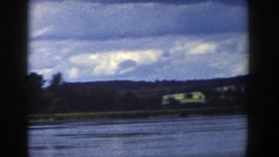 1958 Wisconsin 8mm frame showing calm lake water, a wooded shoreline, distant hills, fields, and a layered cloudy sky.