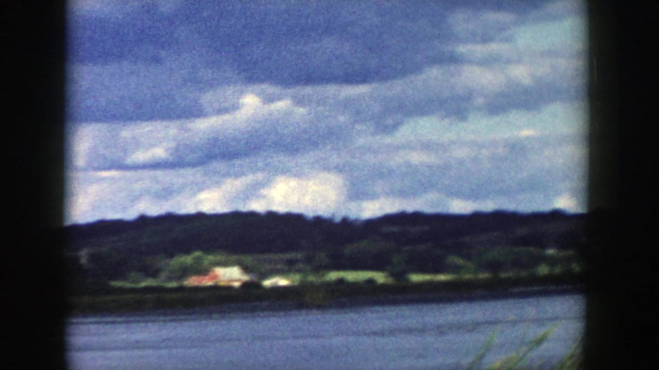 1958 Wisconsin lakeside frame with calm water, dark hills, green fields, a small pale structure, and textured cloud layers.