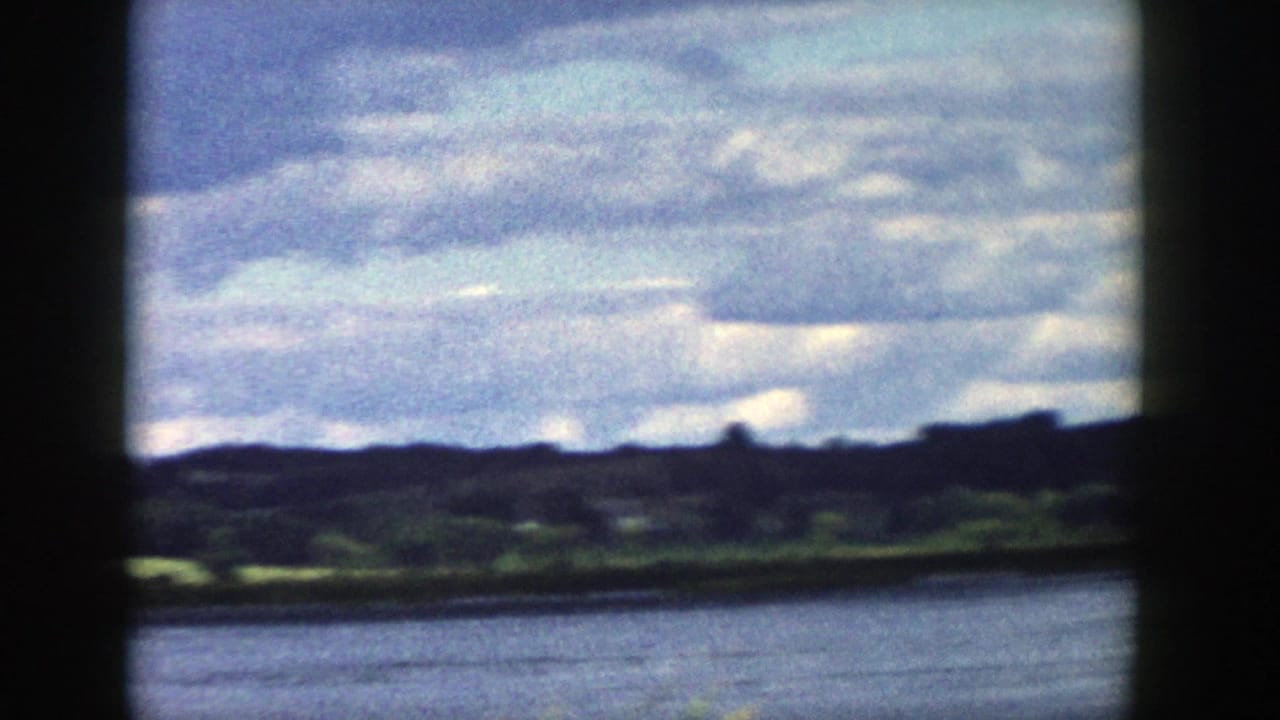 1958 Wisconsin home-movie frame looking across water toward low green hills under a cloudy sky, partly blocked by a dark window frame.