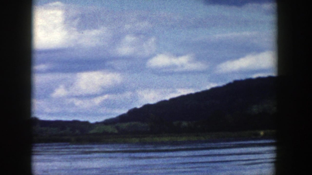 1958 Wisconsin scenic frame showing water, a dark wooded hill, and layered clouds from a moving window viewpoint.