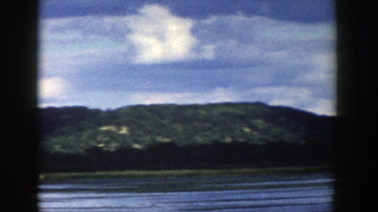 1958 Wisconsin home-movie still of a green hillside across water, with cloudy sky and a dark vertical window bar in the foreground.