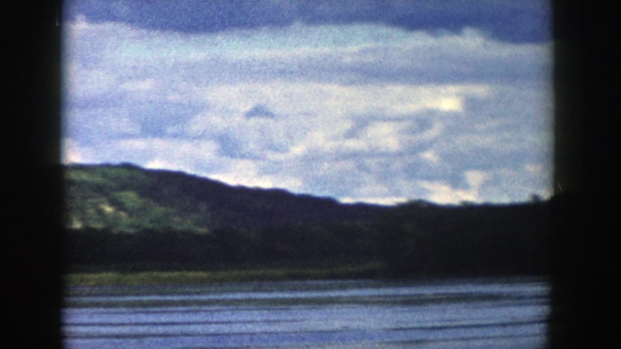 1958 Wisconsin home-movie frame showing a blue lake, dark forested shoreline, low hills, and a cloudy sky.