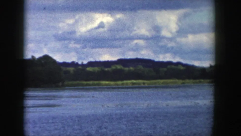 1958 Wisconsin lake view with rippled blue water, green shoreline, low hills, and bright cloud cover.