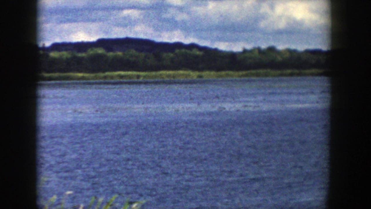 1958 Wisconsin home-movie view across a blue lake toward a tree-lined shore and low dark hill.