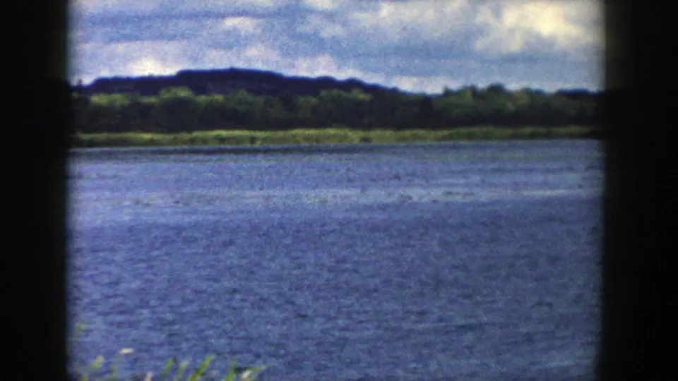 1958 Wisconsin home-movie view across a blue lake toward a tree-lined shore and low dark hill.