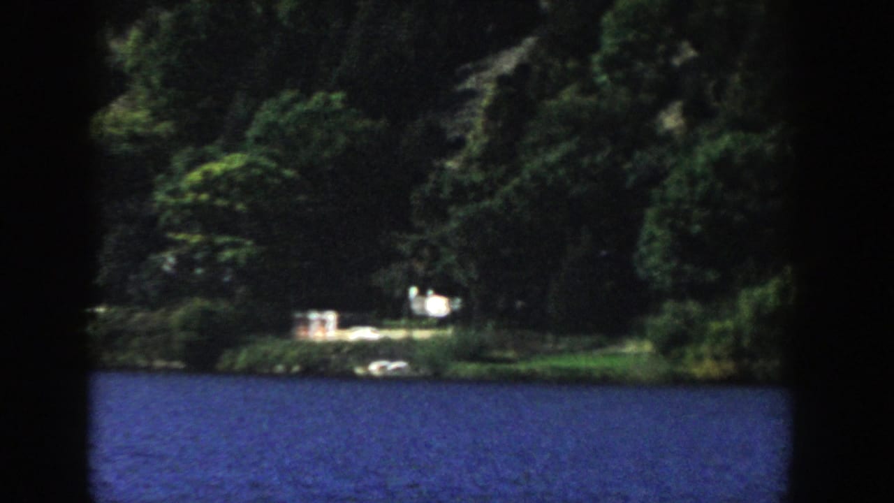 Distant Wisconsin lakeshore with blue water, thick trees, and small light-toned shoreline structures visible through soft 8mm focus.