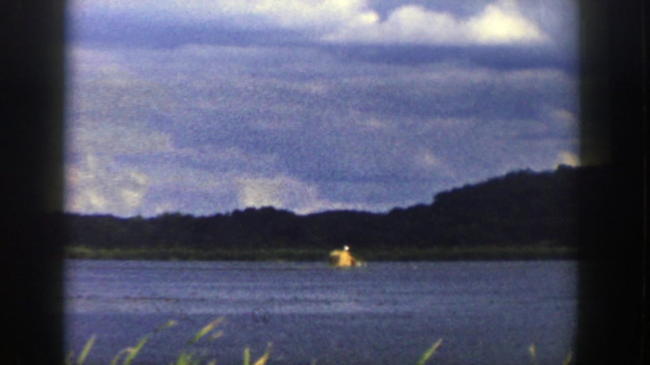 1958 Wisconsin river landscape viewed from a moving train window, with wooded hills, cloudy sky, and a small yellow object on the water.