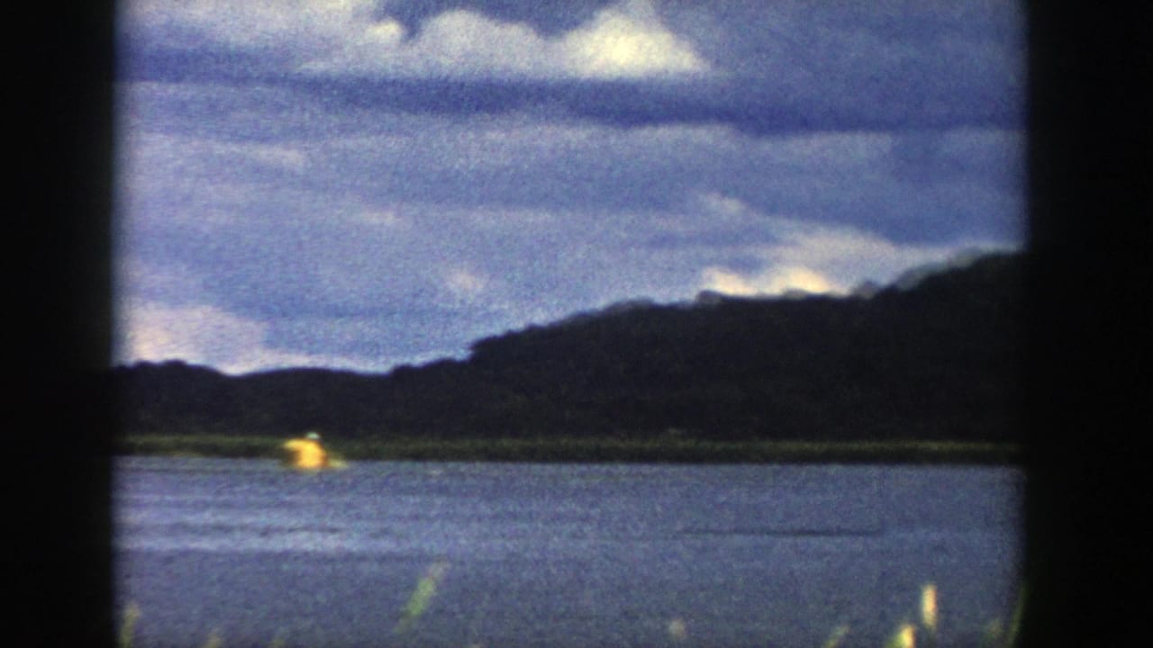 Train-window view in Wisconsin, 1958, showing a blue river, dark wooded hillside, bright clouds, and a small yellow object near the far bank.
