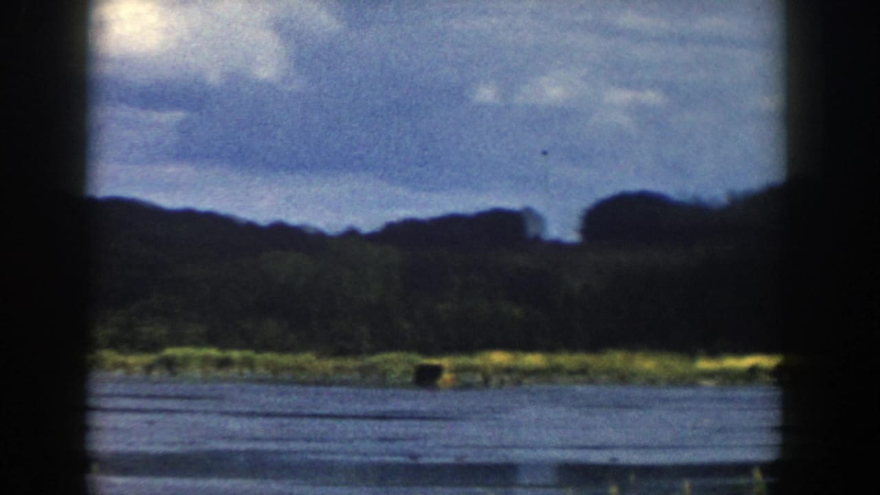Mid-pan 1958 Wisconsin 8mm view of blue-gray water, low green shoreline, distant trees, and cloud cover.