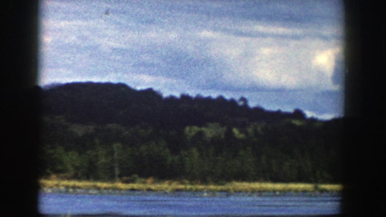 1958 Wisconsin 8mm frame showing calm water, a low wooded shore, distant hills, and overcast sky within dark film borders.