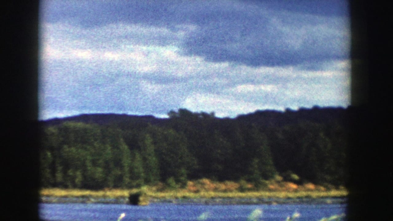 1958 Wisconsin 8mm frame with water, a thin shoreline, dense trees, distant hills, and layered gray clouds.