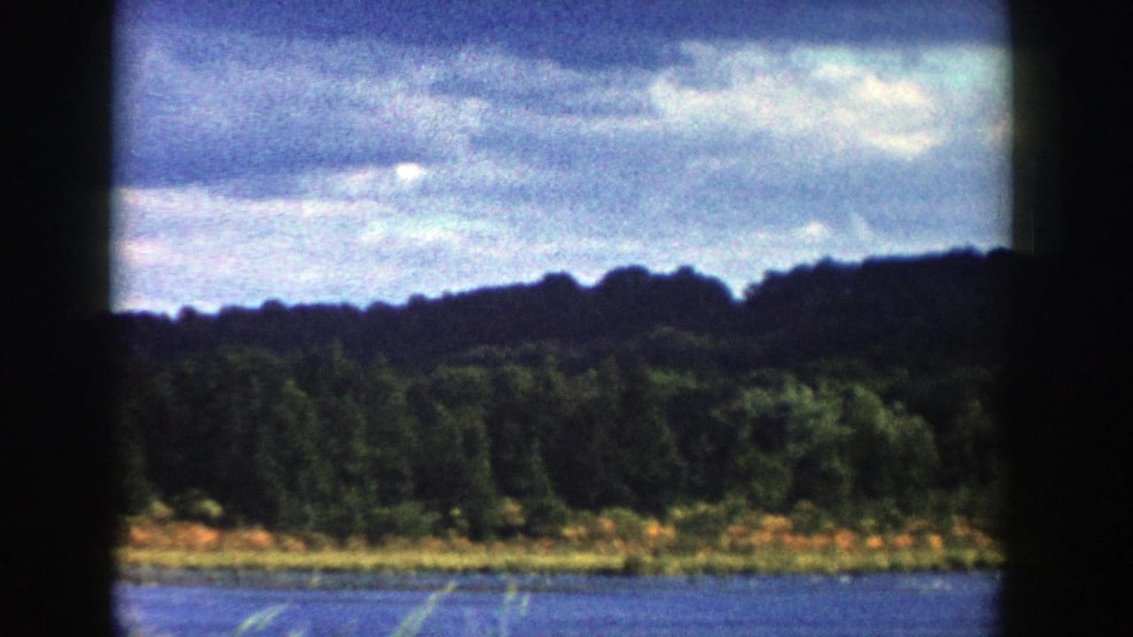 1958 Wisconsin home-movie frame looking over water toward a wooded shore, rolling hills, and a cloudy sky.