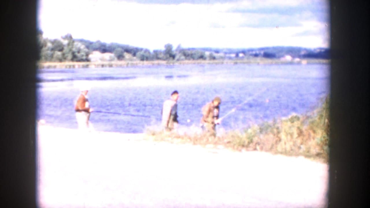 1958 Wisconsin 8mm frame showing two adult men at a bright riverbank, one appearing to hold a fishing rod, with water and distant hills beyond.