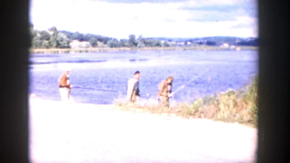 1958 Wisconsin 8mm frame showing two adult men at a bright riverbank, one appearing to hold a fishing rod, with water and distant hills beyond.
