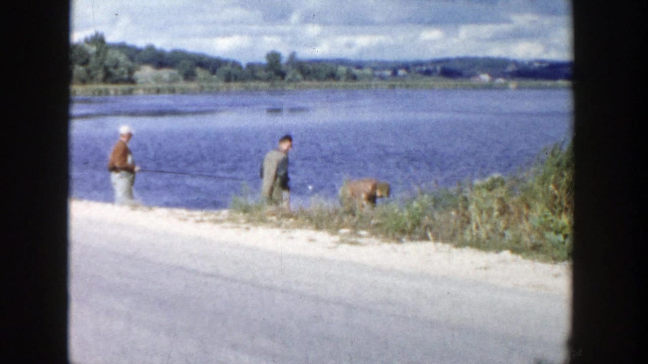 1958 Wisconsin home-movie frame of two adult men and a dog beside a river, with one man fishing from the shoreline.