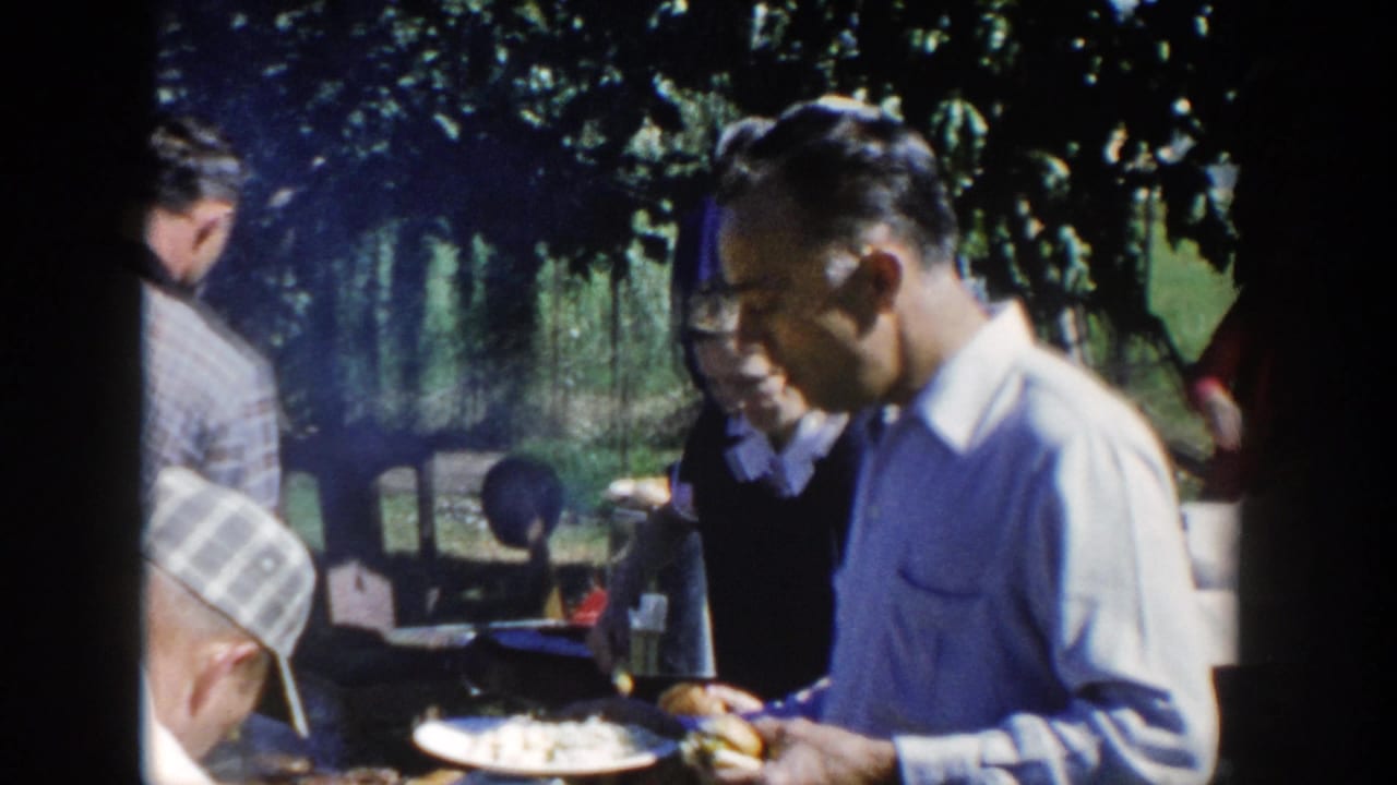1958 Wisconsin 8mm frame showing adults gathered outdoors near a grill, with a man in a light shirt holding a plate in a shaded garden.