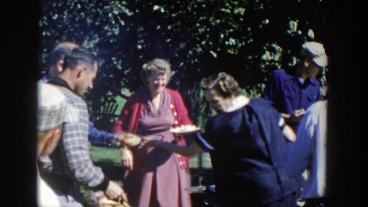 1958 Wisconsin home-movie frame of adults at a backyard barbecue, including a woman in a red dress and others serving food near trees.