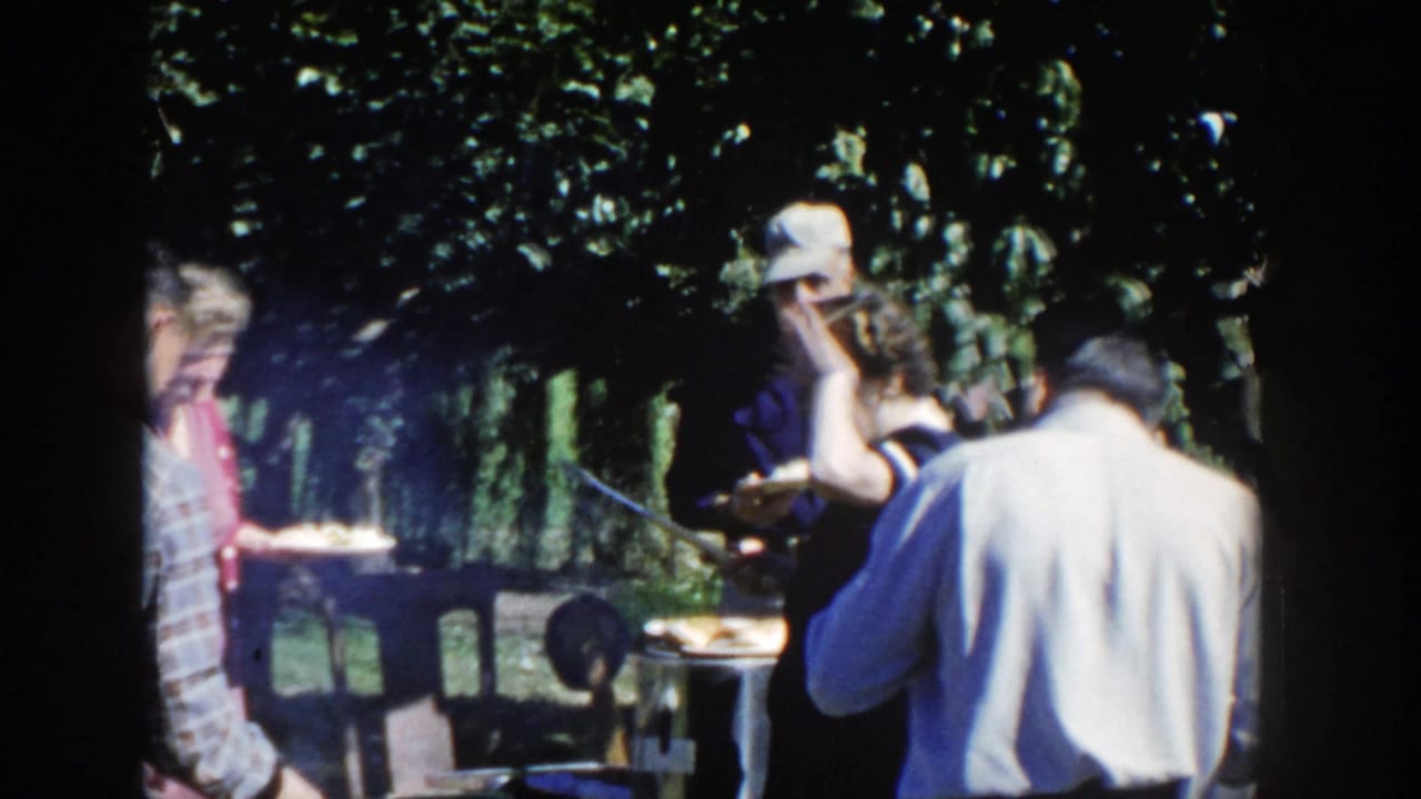 1958 Wisconsin 8mm frame showing several adults near a backyard grill and table, partly shaded by trees during a casual cookout.