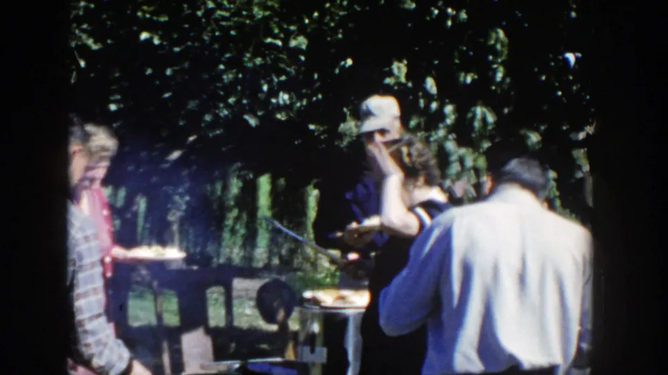 1958 Wisconsin 8mm frame showing several adults near a backyard grill and table, partly shaded by trees during a casual cookout.