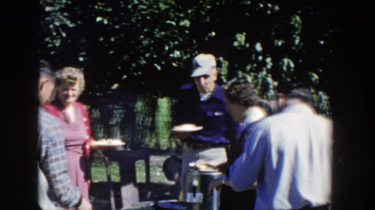 1958 Wisconsin home-movie frame of adults holding plates around a grill, with leafy trees and lawn behind them.
