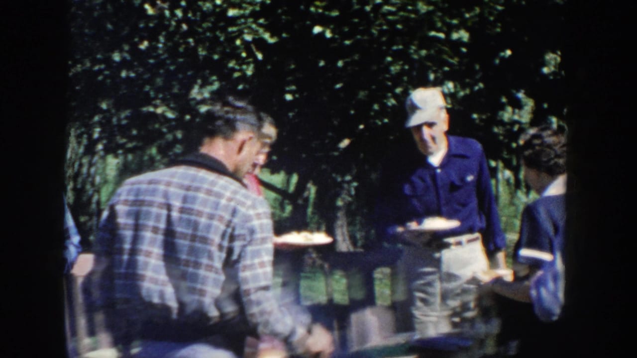 1958 Wisconsin home-movie frame of adults in casual clothing standing and sitting near a shaded outdoor lunch table.