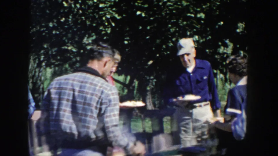 1958 Wisconsin home-movie frame of adults in casual clothing standing and sitting near a shaded outdoor lunch table.
