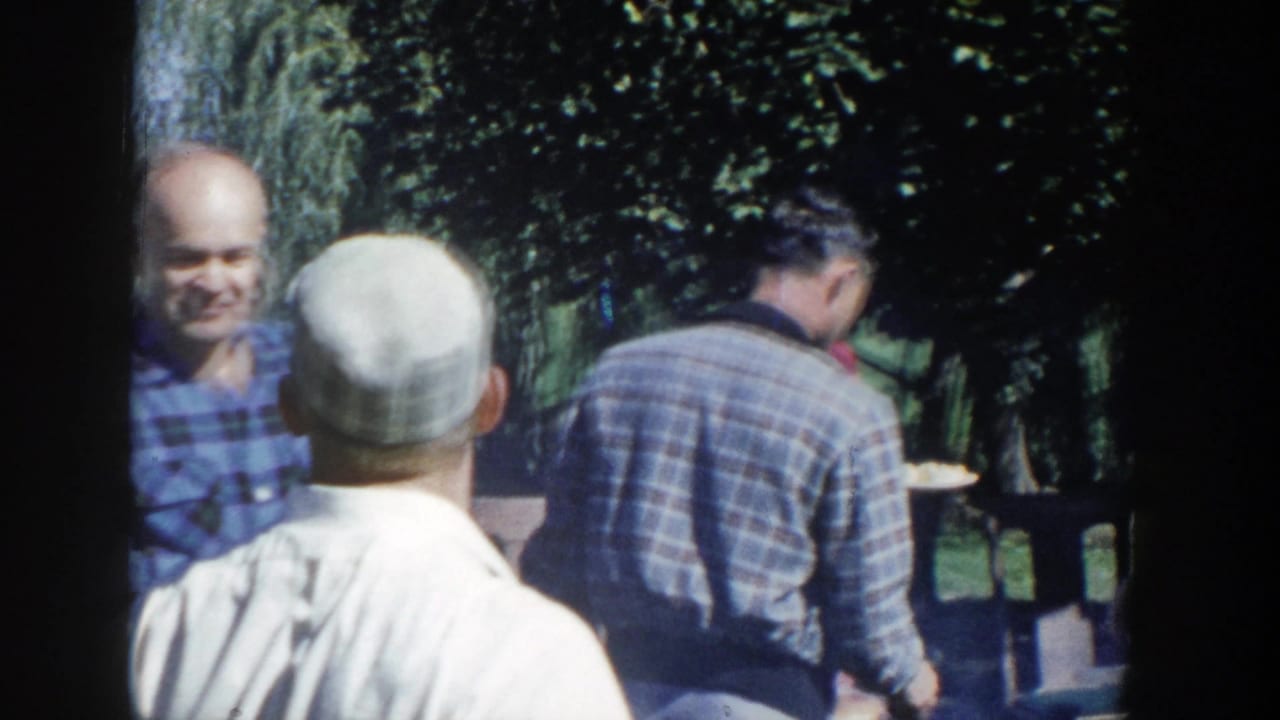 Adults at a 1958 Wisconsin outdoor gathering hold plates and talk beneath trees in a home-movie lunch scene.