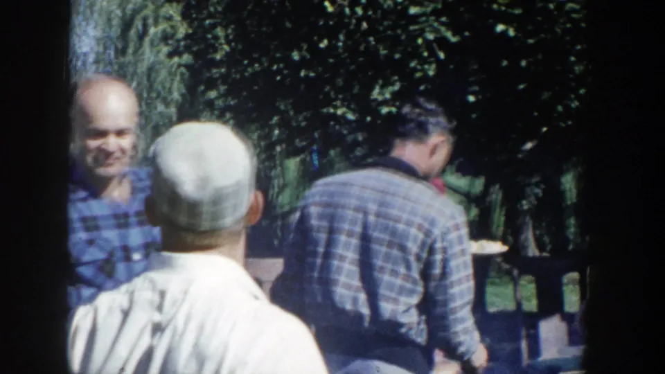 Adults at a 1958 Wisconsin outdoor gathering hold plates and talk beneath trees in a home-movie lunch scene.
