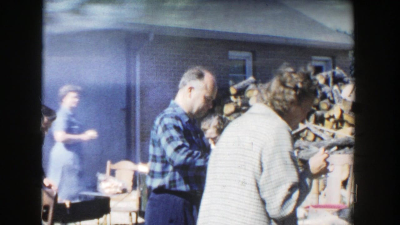 Home-movie frame from Wisconsin in 1958 showing adults eating or serving food beside smoke, firewood, and a building wall.