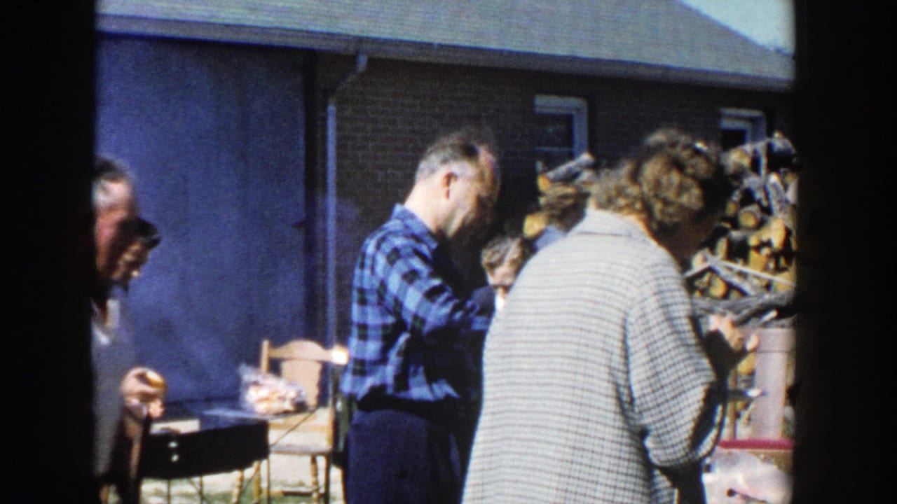 1958 Wisconsin home-movie frame of adults beside a smoky lunch area, with firewood and a building visible behind them.