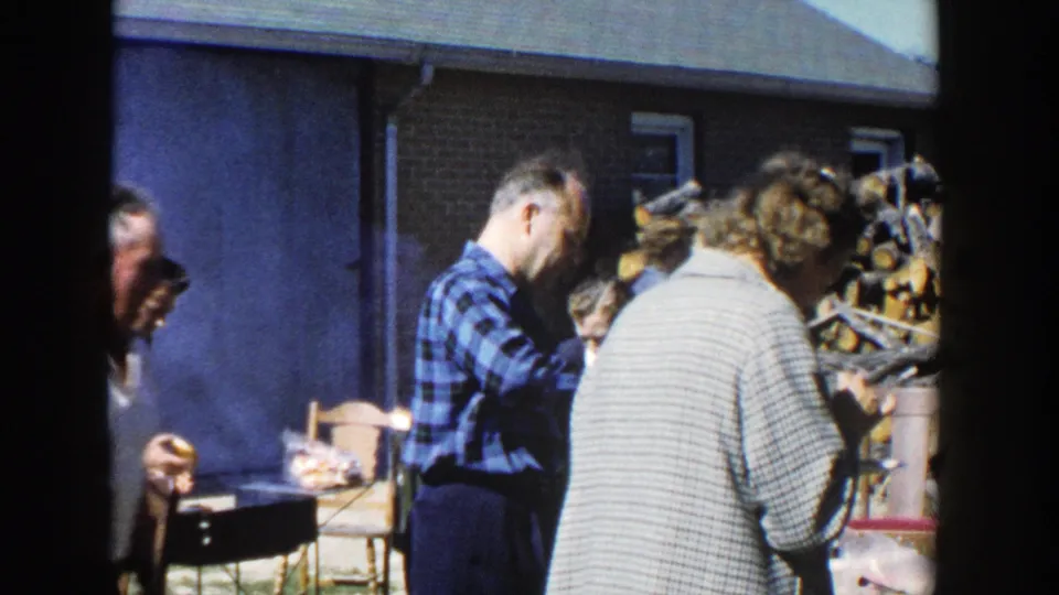 1958 Wisconsin home-movie frame of adults beside a smoky lunch area, with firewood and a building visible behind them.