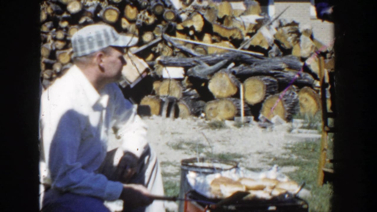 Man in plaid cap sits by an outdoor grill with stacked firewood behind him.