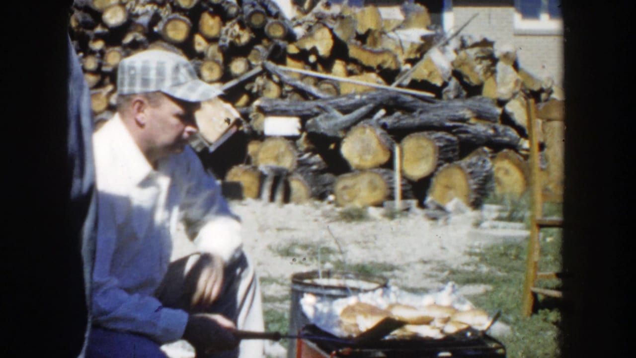 A man in a plaid cap is seated near a portable backyard grill, with chopped logs and yard objects visible behind him.