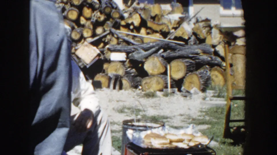 A standing adult passes close to the camera, partly blocking the view of the seated man, backyard grill, and stacked firewood.