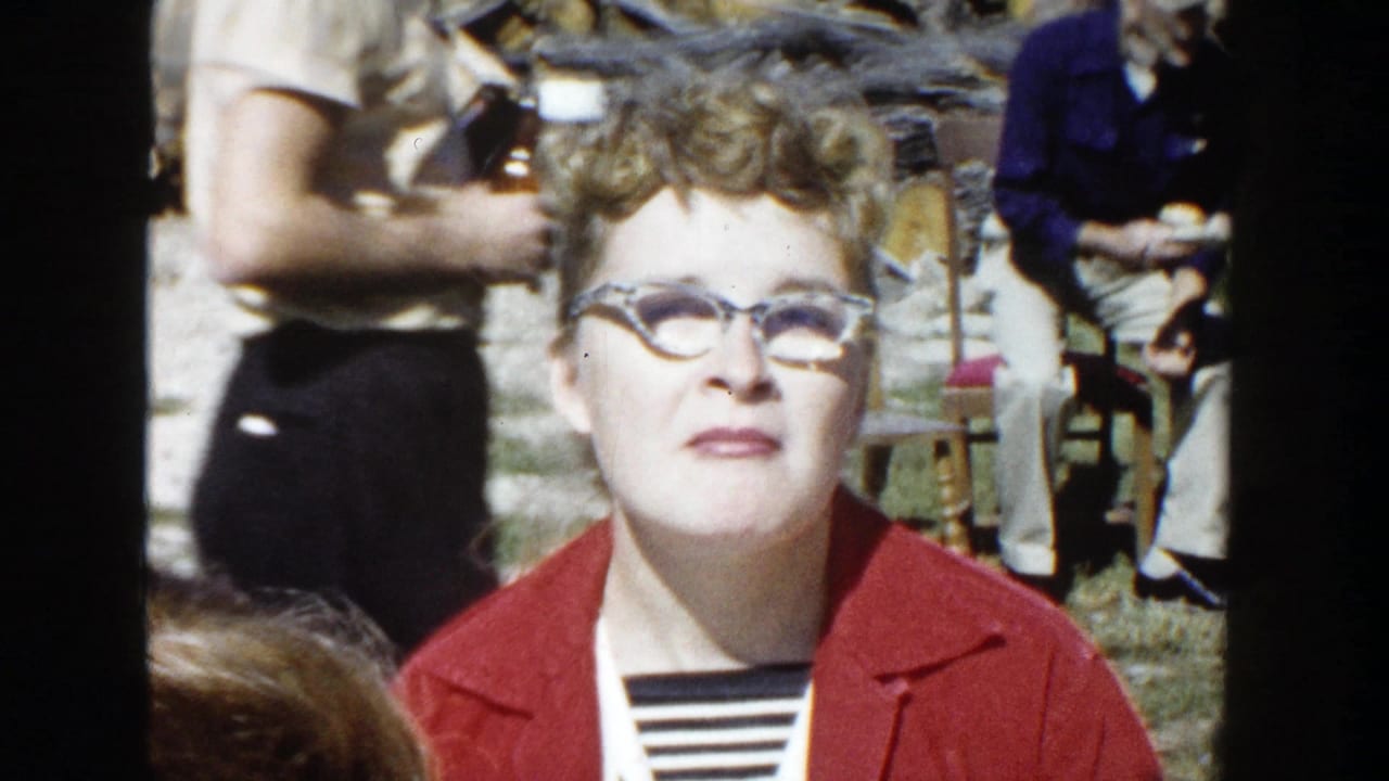 In 1958 Wisconsin home-movie footage, a woman with glasses and a red jacket looks toward the camera in a sunny yard.