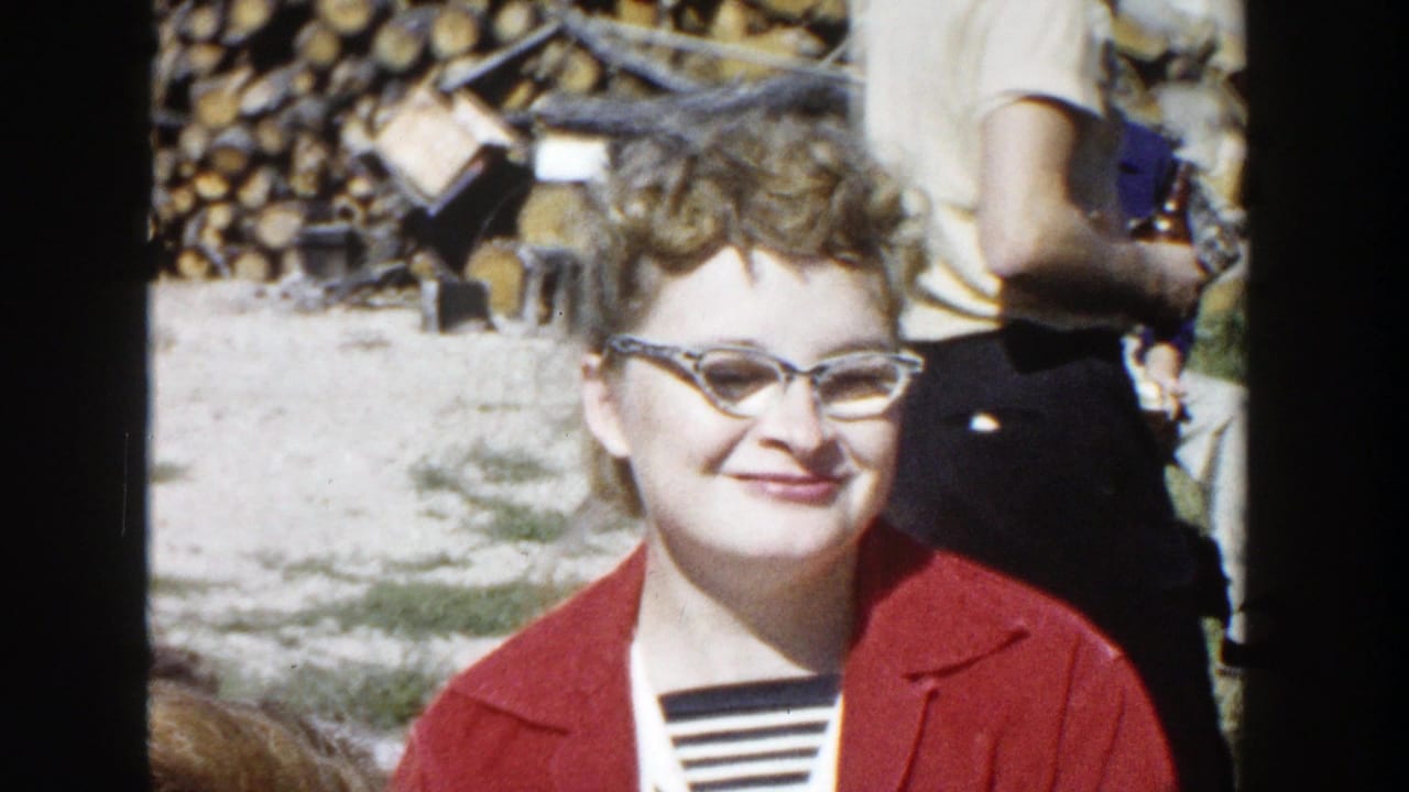 A woman in glasses and a red jacket smiles faintly toward the camera in a 1958 Wisconsin outdoor home-movie scene.